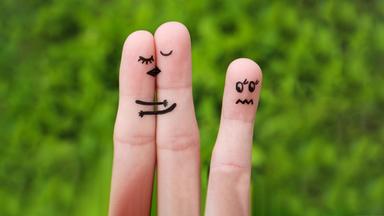 zwei Finger schmiegen sich aneinander, auf Ihnen die Gesichtsmimik von sich Küssenden aufgemalt, ein einzelner Finger mit aufgemalter unglücklicher Mimik im Abseits