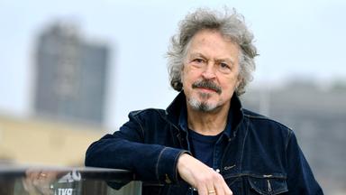 Ein Mann mit grauen lockigen Haaren stützt sich auf ein Geländer.