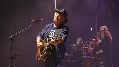 Ein älterer Mann mit Bart und Hut spielt Gitarre auf einer Bühne und lächelt.