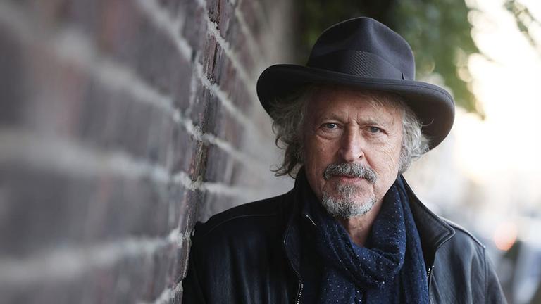 Mann mit Hut steht an einer Mauer und schaut in die Kamera