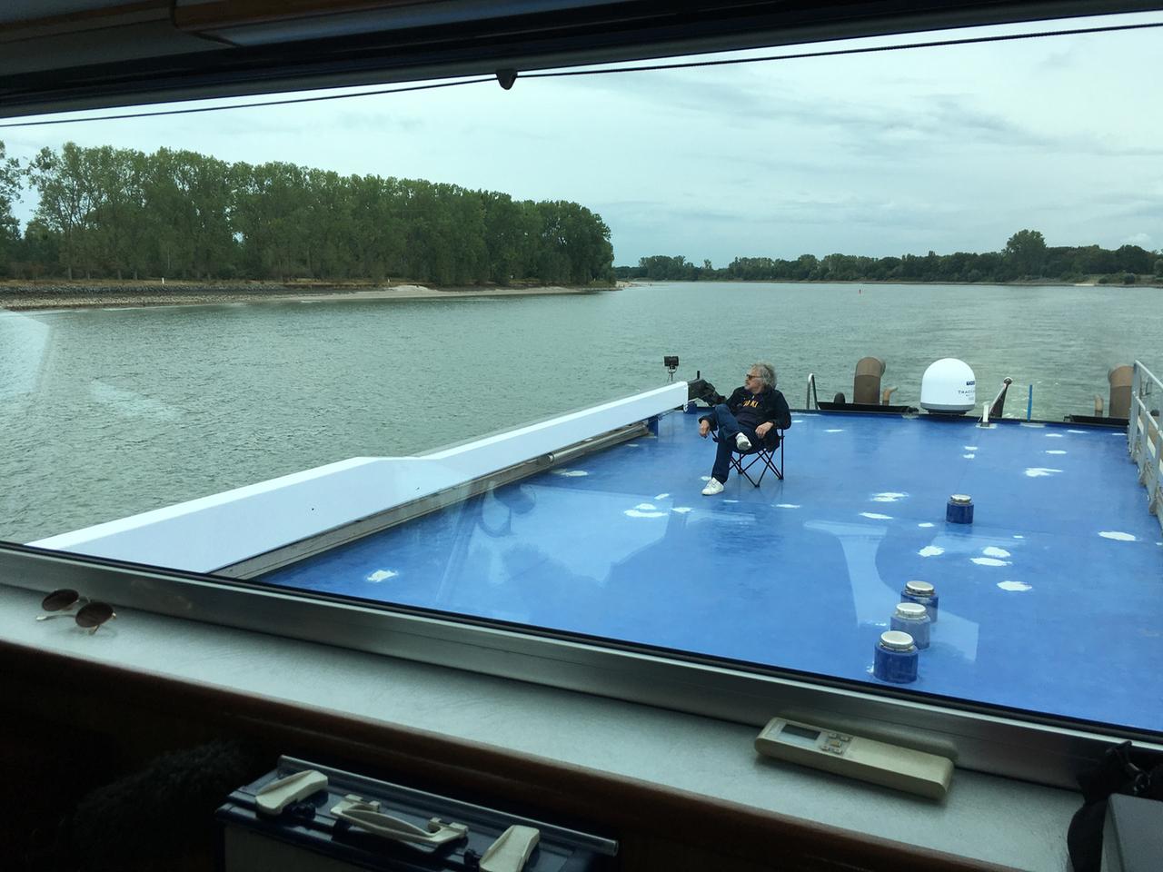 Wolfgang Niedecken sitzt an Deck des Schubleichters in einem Klappstuhl und schaut auf den Rhein und genießt die Landschaft.