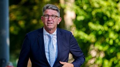 Ein Mann mit graumelierten kurzen Haaren, Brille und im dunkelblauen Anzug mit hellblauem Hemd und Krawatte fasst sich im Laufen mit der linken Hand vorne ans Jackett.