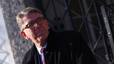 Ein Mann mit graumelierten kurzen Haaren, Brille und im dunklen Anzug und Mantel schaut nach links.