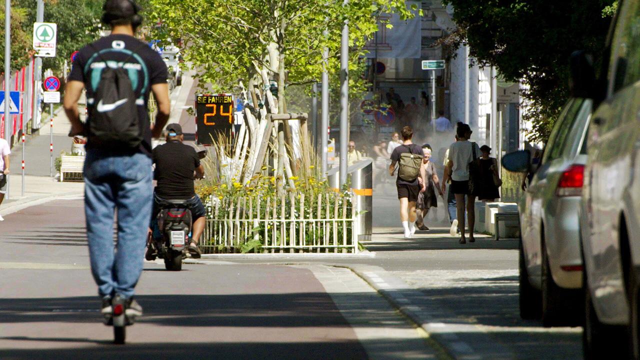Menschen, Fahrzeuge und Autos auf einer Straße mit Geschwindigkeitsanzeige.