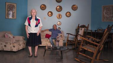 Das Bild zeigt eine Wohnzimmer-Szene. In der Mitte ist eine ältere Frau auf der linken Seite des Rahmens zu sehen. Sie trägt ein kurzärmliges, buntes Oberteil mit einer weißen Grundfarbe und einem blau-roten Muster sowie einen dunklen, knielangen Rock. Die Frau hat graue Haare, die zu einem Dutt hochgesteckt sind.   Auf der rechten Seite sitzt ein älterer Mann auf einer Couch, die mit einem hellbeigen Stoff bezogen ist. Er trägt ein kariertes Hemd und eine graue Hose. Hinter ihm befindet sich ein Kissen in einem warmen, rosafarbenen Farbton.   Im Hintergrund sind mehrere dekorative Wandplatten unterschiedlichster Motive angebracht. Die Wände sind in einem hellblauen Farbton gestrichen. Der Boden ist mit einem grauen, glatten Material ausgelegt, das leicht glänzt. Auf einem niedrigen Tisch in der Mitte der Szene steht eine Blumenvase, umgeben von einigen weiteren kleinen Objekten.  Das gesamte Ambiente wirkt heimelig und zeigt Elemente des Alltagslebens.