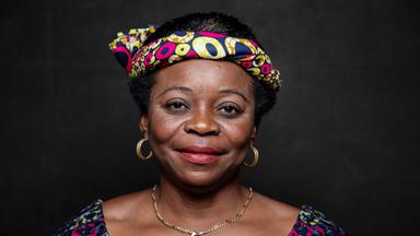 Das Bild zeigt eine Frau mit dunkler Hautfarbe und afrokrausem Haar. Sie trägt ein farbenfrohes Kopftuch, das mit einem Muster aus vielen bunten Punkten und Formen verziert ist. Ihre Oberbekleidung hat ein ähnliches buntes Muster und wirkt traditionell. Sie hat große, goldene Ohrringe und eine goldene Halskette. Ihr Gesicht ist freundlich und strahlt Selbstbewusstsein aus. Der Hintergrund ist dunkel und sorgt dafür, dass die Frau stark im Vordergrund steht.   Die Bildunterschrift erwähnt, dass es sich um eine Aufnahme aus dem Film "Woman" handelt, der Frauen und ihre Geschichten aus der ganzen Welt thematisiert.