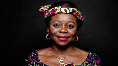 Das Bild zeigt eine Frau mit dunkler Hautfarbe und afrokrausem Haar. Sie trägt ein farbenfrohes Kopftuch, das mit einem Muster aus vielen bunten Punkten und Formen verziert ist. Ihre Oberbekleidung hat ein ähnliches buntes Muster und wirkt traditionell. Sie hat große, goldene Ohrringe und eine goldene Halskette. Ihr Gesicht ist freundlich und strahlt Selbstbewusstsein aus. Der Hintergrund ist dunkel und sorgt dafür, dass die Frau stark im Vordergrund steht.   Die Bildunterschrift erwähnt, dass es sich um eine Aufnahme aus dem Film "Woman" handelt, der Frauen und ihre Geschichten aus der ganzen Welt thematisiert.