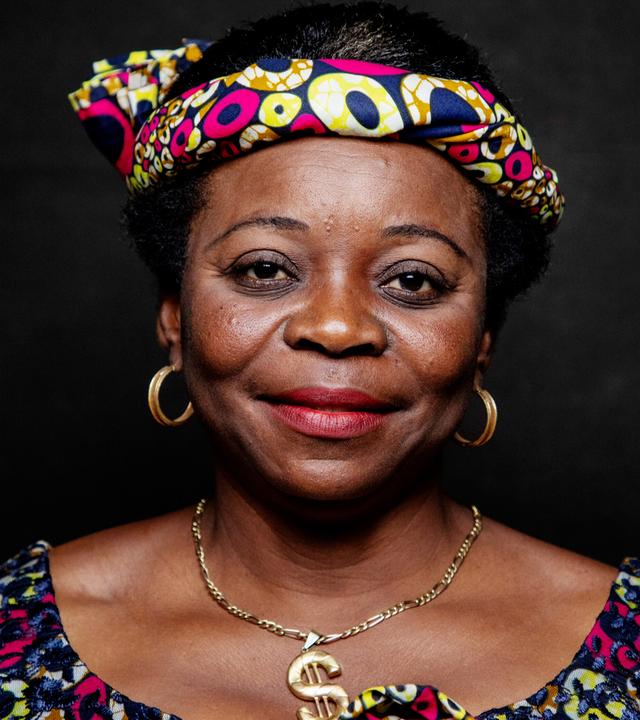 Das Bild zeigt eine Frau mit dunkler Hautfarbe und afrokrausem Haar. Sie trägt ein farbenfrohes Kopftuch, das mit einem Muster aus vielen bunten Punkten und Formen verziert ist. Ihre Oberbekleidung hat ein ähnliches buntes Muster und wirkt traditionell. Sie hat große, goldene Ohrringe und eine goldene Halskette. Ihr Gesicht ist freundlich und strahlt Selbstbewusstsein aus. Der Hintergrund ist dunkel und sorgt dafür, dass die Frau stark im Vordergrund steht.   Die Bildunterschrift erwähnt, dass es sich um eine Aufnahme aus dem Film "Woman" handelt, der Frauen und ihre Geschichten aus der ganzen Welt thematisiert.