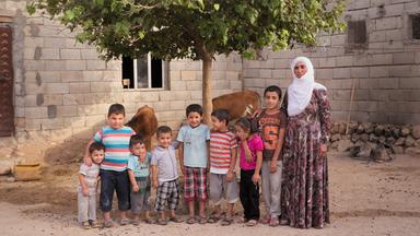 Das Bild zeigt eine Gruppe von insgesamt neun Kindern und einer Frau, die in einem ländlichen Umfeld steht. Die Szene ist vor einem einfachen, aus grauen Steinen gebauten Haus platziert. Hinter der Gruppe befinden sich zwei braune Kühe, die ebenfalls im Bild sichtbar sind, und ein kleiner Baum spendet etwas Schatten.  Die Frau trägt ein langes, gemustertes Kleid sowie ein Kopftuch, was auf einen traditionellen Kleidungsstil hinweist. Sie steht auf der rechten Seite des Bildes und hat einen freundlichen Gesichtsausdruck. Um sie herum stehen die Kinder, die in verschiedenen Altersgruppen zu sehen sind. Einige Kinder tragen gestreifte T-Shirts, während andere bunte oder einfarbige Kleidung tragen. Ihr Gesichtsausdruck zeigt eine Mischung aus Neugier und Freude.   Das Umfeld ist unbefestigt, und es sind einige Steine und Erde zu sehen. Die Lichtverhältnisse scheinen warm und einladend zu sein, was eine positive und harmonische Atmosphäre vermittelt.