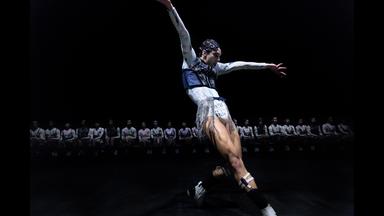 Im Vordergrund sieht man eine Person, die tanzt und im Hintergrund sitzen viele Menschen in einer Reihe.