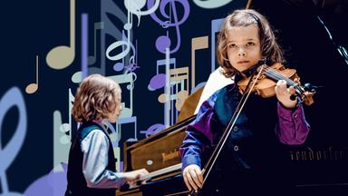 Zwei musizierende Kinder. Der Junge rechts hält eine Geige, der Junge links hinten spielt am Klavier.