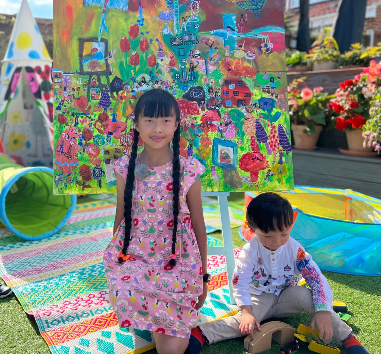 Ein Mädchen mit langen braunen Zöpfen steht draußen im Garten vor einem bunten Gemälde. Neben ihr auf dem Boden sitzt ein kleiner Junge, der mit einer Holzeisenbahn spielt.