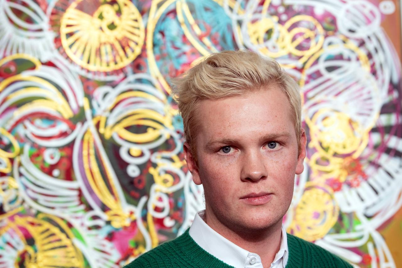 Großaufnahme eines jungen Mannes mit blonden, kurzen Haaren vor einem abstraktenn, buntem Gemälde