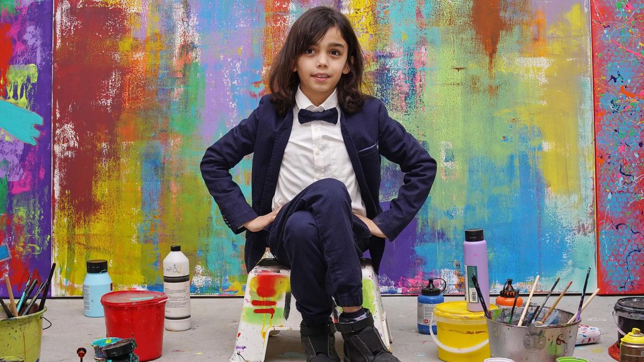 Ein Jungte mit langen Haaren, Anzugsjacke, weißem Hemd und schwarzer Fliege vor einem abstrakten Gemälde. Neben ihm auf dem Boden Farbeimer und Pinsel.