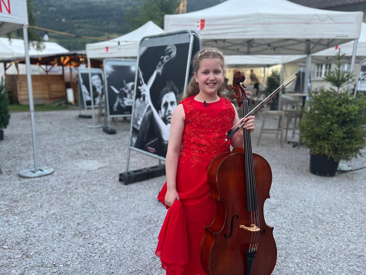 Mädchen in rotem Kleid hält Cello und Bogen in der Hand und steht lächelnd vor einem SW-Musik-Plakat und weißen Pavillons. Mit der rechten Hand hält sie ihr leicht gerafftes Kleid hoch.