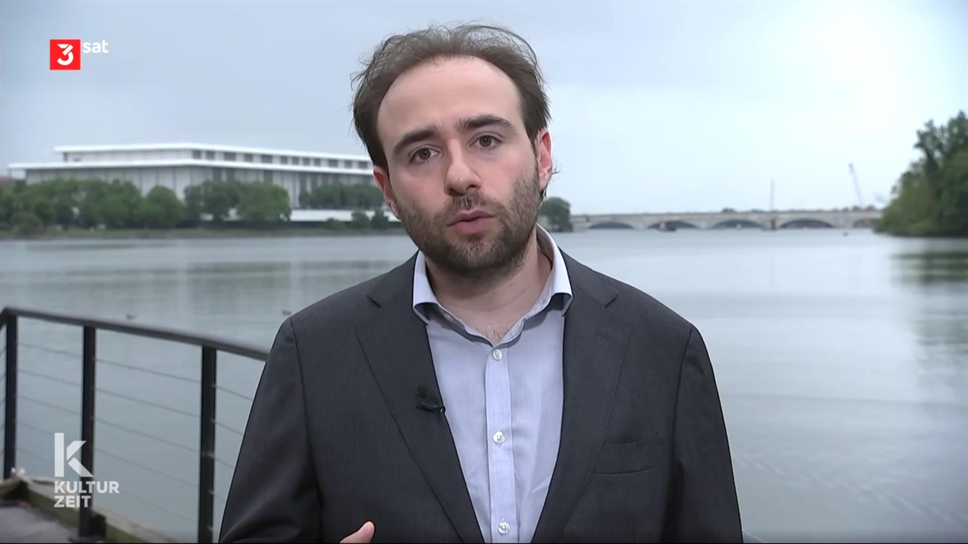 Bild eines Mannes, der in die Kamera schaut, im Hintergrund ist ein Fluss mit einem großen Gebäude am Ufer zu sehen.