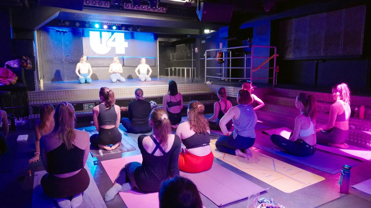 Eine Gruppe von Menschen praktiziert Yoga in einem Club, im Hintergrund eine Bühne mit U4-Logo.