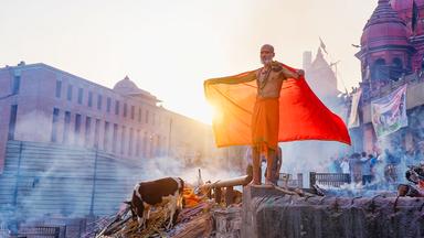 Ein Mann mit ausgebreitetem orangen Tuch auf einer Mauer stehend, neben einem lodernden Scheiterhaufen auf dem sich eine Kuh befindet.