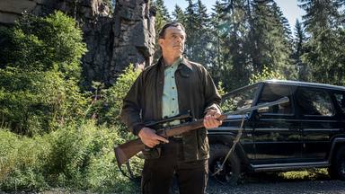 Ein älterer Mann mit Jagdgewehr steht vor einem Felsen und einem schwarzen Geländewagen im Wald.