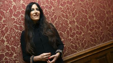 Eine Frau mit langem braunen Haar und einer schwarzen Bluse steht vor einer rot gemusterten Tapete.