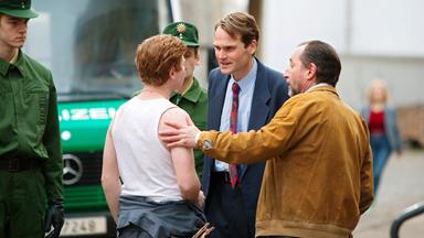 Zwei Männer in Anzug und Lederjacke unterhalten sich mit einem rothaarigen jüngeren Mann in einem weißen Unterhemd. Zwei Polizisten in Uniform beobachten die drei Männer.