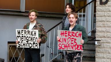 Zwei junge Männer und eine junge Frau stehen an der Treppe eines Gebäudes. Die beiden Männer halten jeweils ein Protestplakat vor sich.