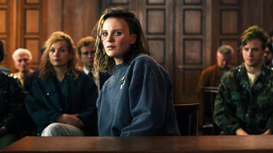 Eine junge Frau sitzt an einem Tisch in einem Saal. Hinter ihr sitzen Leute im Saal.