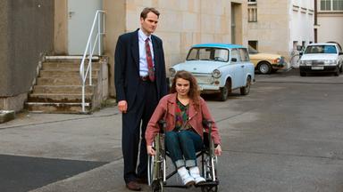 Ein Mann im Anzug steht neben einer Frau im Rollstuhl. Beide befinden sich in einem Hinterhof,.