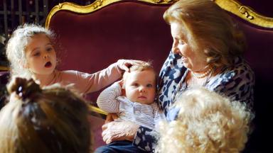 Auf dem Bild sieht man eine sehr alte Frau auf einen edlen, antiken Sofa mit Goldrand sitzen. Sie hält ein Kleinkind auf dem Arm, neben ihr sitzt ein etwa 3-jähiges Mädchen, das dem Kleinkind über den Kopf streichelt. Die alte Frau trägt feine Kleidung und Ohrringe und lächelt das Kleinkind an. Vor dem Sofa sieht man ein weiteres, blondgelocktes Kind von hinten, daneben, ebenfalls von hinten eine Frau mit braunen Haaren und Dutt.