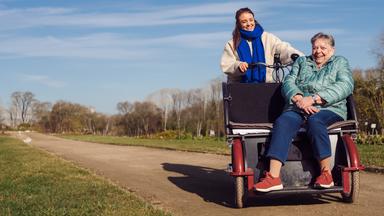 Junge Frau steht neben ihrer älteren Freundin, die im Rollstuhl sitzt.
