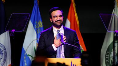 Ein Mann mit Bart und im Anzug steht hinter einem Rednerpult vor Flaggen und legt sich lächelnd eine Hand aufs Herz