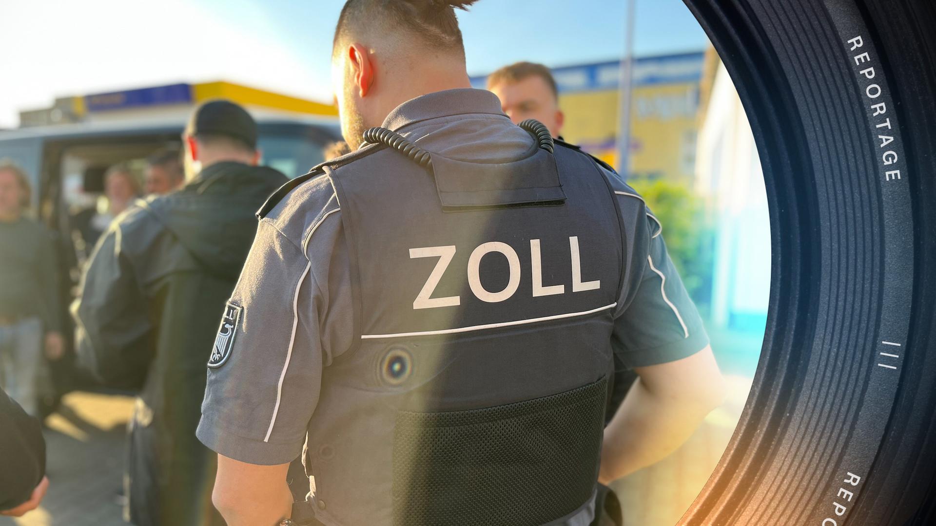 Eine Gruppe Zollbeamten und Zollbeamtinnen steht im Freien. Zentral steht ein Beamter mit dem Rücken zur Kamera. Seine Stichschutzweste trägt den Schriftzug "Zoll".