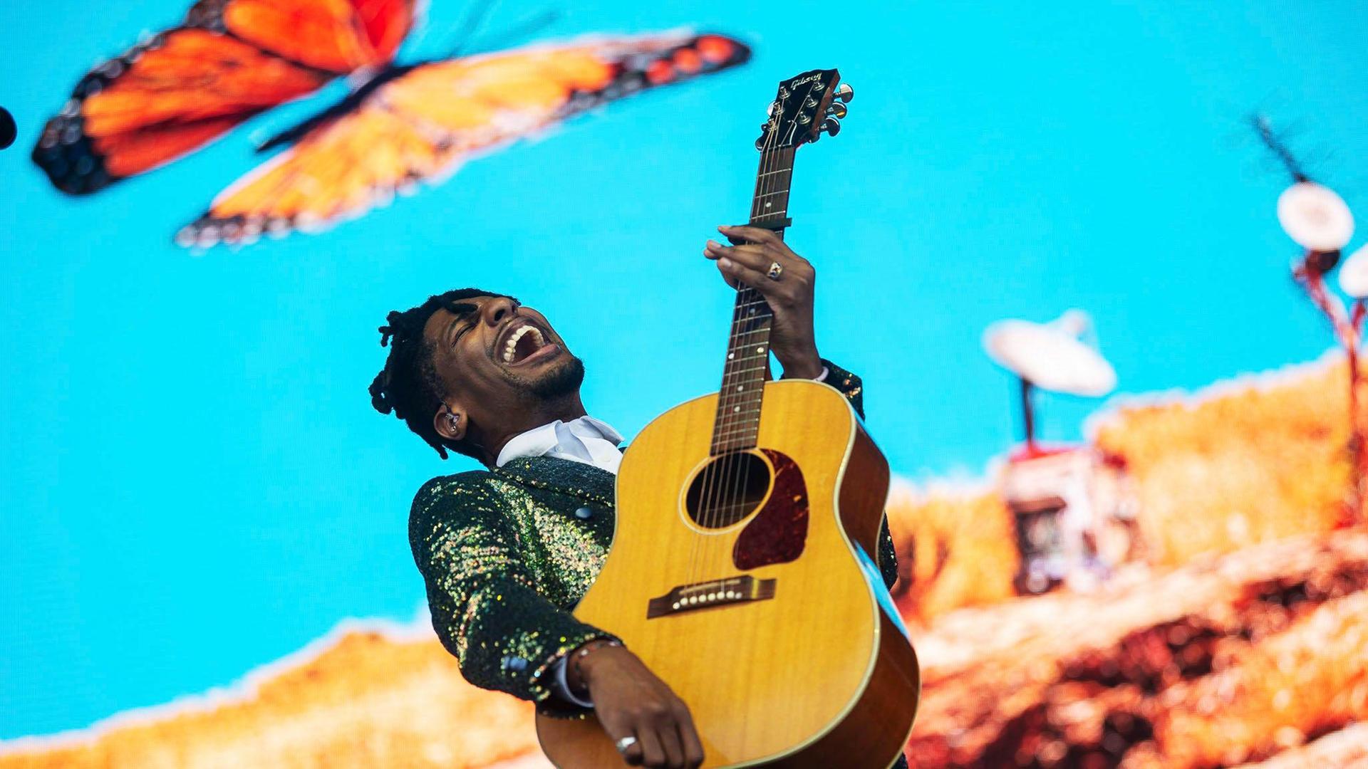 Ein Mann mit Gitarre in der Hand singt vor hellblauem Hintergrund. Links über ihm sieht man einen großen Schmetterling.