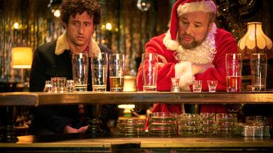 Zwei Männer sitzen an einem Tresen in einer Bar; der eine trägt ein Weihnachtsmannkostüm, der andere eine Jacke mit Fellkragen. Vor ihnen stehen zahlreiche leere Bier- und Schnapsgläser.