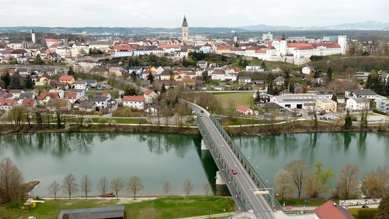Das Bild zeigt eine Landschaft mit einem Fluss, der sich durch eine Stadt windet. Im Vordergrund ist eine Brücke zu sehen, die über den Fluss führt. Auf der Brücke sind mehrere Autos unterwegs. Auf der linken Seite des Bildes befinden sich Bäume und eine kleine Wiese.   Im Hintergrund erhebt sich die Stadt mit vielen Gebäuden, die entweder mehrstöckig oder einstöckig sind, in verschiedenen Farben, hauptsächlich beige, rot und weiß. Ein hervorgehobenes Gebäude mit einem hohen Turm ist offensichtlich das Stadtzentrum oder eine Kirche. Die Stadtsilhouette ist überwiegend flach, während im Hintergrund sanfte Hügel oder Berge zu erkennen sind.   Der Himmel ist leicht bewölkt, und die gesamte Szene vermittelt den Eindruck eines ruhigen Frühlingsnachmittags. Es gibt eine klare Reflexion des Himmels im Wasser des Flusses.