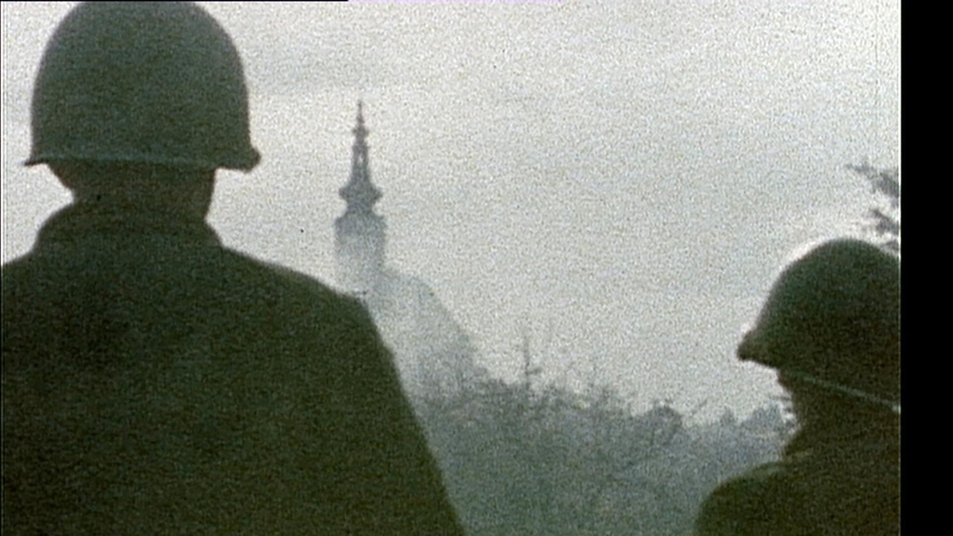 Das Bild zeigt zwei amerikanische Soldaten im Jahr 1945, die in Richtung einer unscharfen Stadtlandschaft blicken. Beide Soldaten tragen helle Stahlhelme und sind von hinten sichtbar. Sie stehen zusammen und blicken auf einen im Hintergrund leicht erkennbaren Turm, der Teil eines Gebäudes ist. Der Himmel über ihnen ist durch einen grauen Schleier verhangen, und die gesamte Szene hat eine melancholische und diffuse Lichtstimmung. Die Umgebung wirkt düster und lässt den Eindruck von Unruhe und Unsicherheit in der Luft entstehen. Es ist klar, dass es sich bei der dargestellten Situation um ein historisches Ereignis zur Zeit des Kriegsendes handelt, wobei die Kulisse vermutlich eine österreichische Stadt im Frühjahr 1945 darstellt.