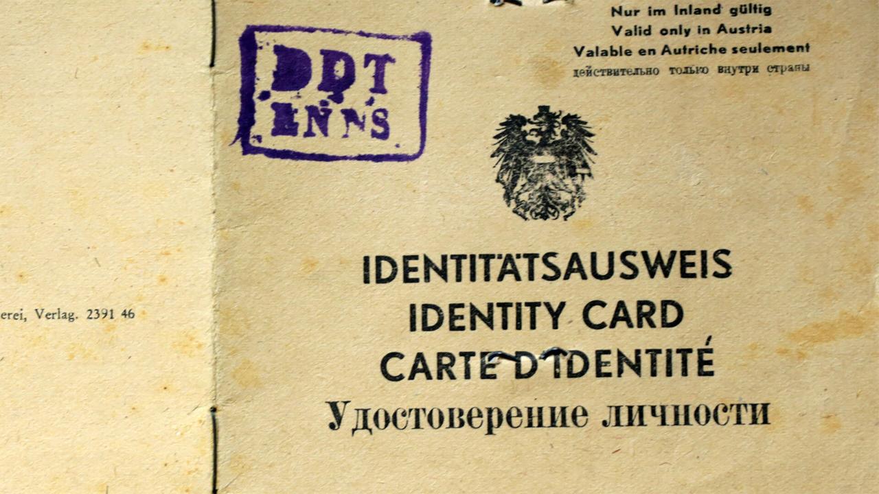 Das Bild zeigt einen alten Identitätsausweis, der auf einem beige Hintergrund liegt. Der Ausweis ist in mehreren Sprachen betitelt: „IDENTITÄTSAUSWEIS“, „IDENTITY CARD“, „CARTE D’IDENTITÉ“ und „Удостоверение личности“, was „Identitätsnachweis“ bedeutet.   Oben links im Ausweis befindet sich ein Stempel, auf dem in lila Farbe die Buchstaben „DPT LNN“ stehen. In der Mitte des Ausweises prangt ein Wappen mit einem stilisierten Adler, ein typisches Symbol für nationale Identifikation in vielen Ländern.   Der Text „Nur im Inland gültig“ und die entsprechenden Übersetzungen weisen darauf hin, dass der Ausweis nur in Österreich anerkannt wird. Der Ausweis hat sichtbare Gebrauchsspuren, wie leichte Flecken und Knicke, die auf sein Alter hinweisen.   Im Hintergrund sind keine weiteren Elemente zu sehen, die auf spezifische Orte oder Ereignisse hinweisen.