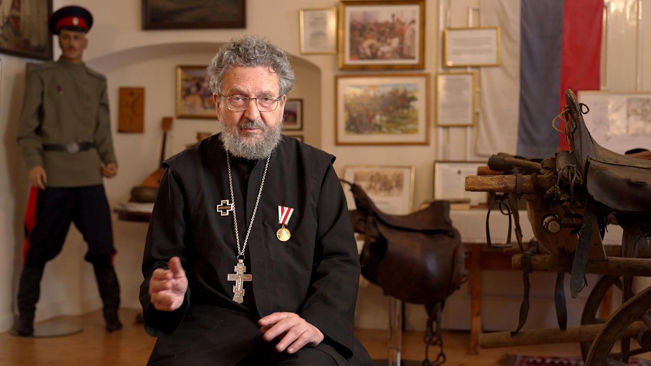 Das Bild zeigt einen älteren Mann mit grauem Haar und einem Bart, der in einem dunklen Gewand sitzt. Er trägt mehrere symbolische Kreuze und eine Auszeichnung um den Hals. Die Person zeigt eine nachdenkliche Miene und gestikuliert mit den Händen.   Im Hintergrund sind verschiedene Gegenstände zu sehen, darunter Bilderrahmen mit Dokumenten und historischen Gemälden sowie eine russische Flagge, die teilweise sichtbar ist. Links neben ihm steht eine lebensgroße Figur in einer historischen Uniform, die an die Zeit der Weißen Armee erinnert. Auf einem Tisch vor ihm sind ein Sattel und andere Objekte aus der Geschichte. Der Raum ist hell beleuchtet und scheint ein Museum oder eine Ausstellung zu sein.