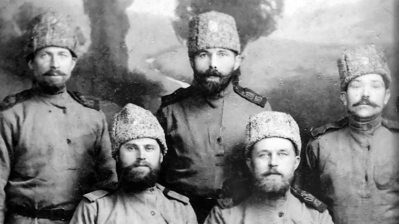 Das Bild zeigt fünf Männer, die in militärischer Kleidung der Weißen Armee aus der Zeit des Russischen Bürgerkriegs (ca. 1917-1923) abgebildet sind. Sie tragen Konsolenjacken und hohe, filzige Hüte mit einem charakteristischen, breiten Rand, die für Kosaken typisch sind. Die Männer stehen in einer Reihe, wobei zwei im Vordergrund sitzen und drei im Hintergrund stehen.   Die Umgebung wirkt unscharf, mit einem unsichtbaren Weg, der hinter ihnen zu erkennen ist, was auf eine ländliche oder natürliche Umgebung hinweist. Das Bild hat einen sepiafarbenen Ton, was ihm ein historisches Aussehen verleiht. Einige Details sind verblasst, was möglicherweise auf das Alter des Fotos hinweist.   Es sind keine spezifischen Gebäude oder andere deutlich erkennbare Attribute im Bild vorhanden, außer der beschriebenen militärischen Bekleidung und der Kulisse.